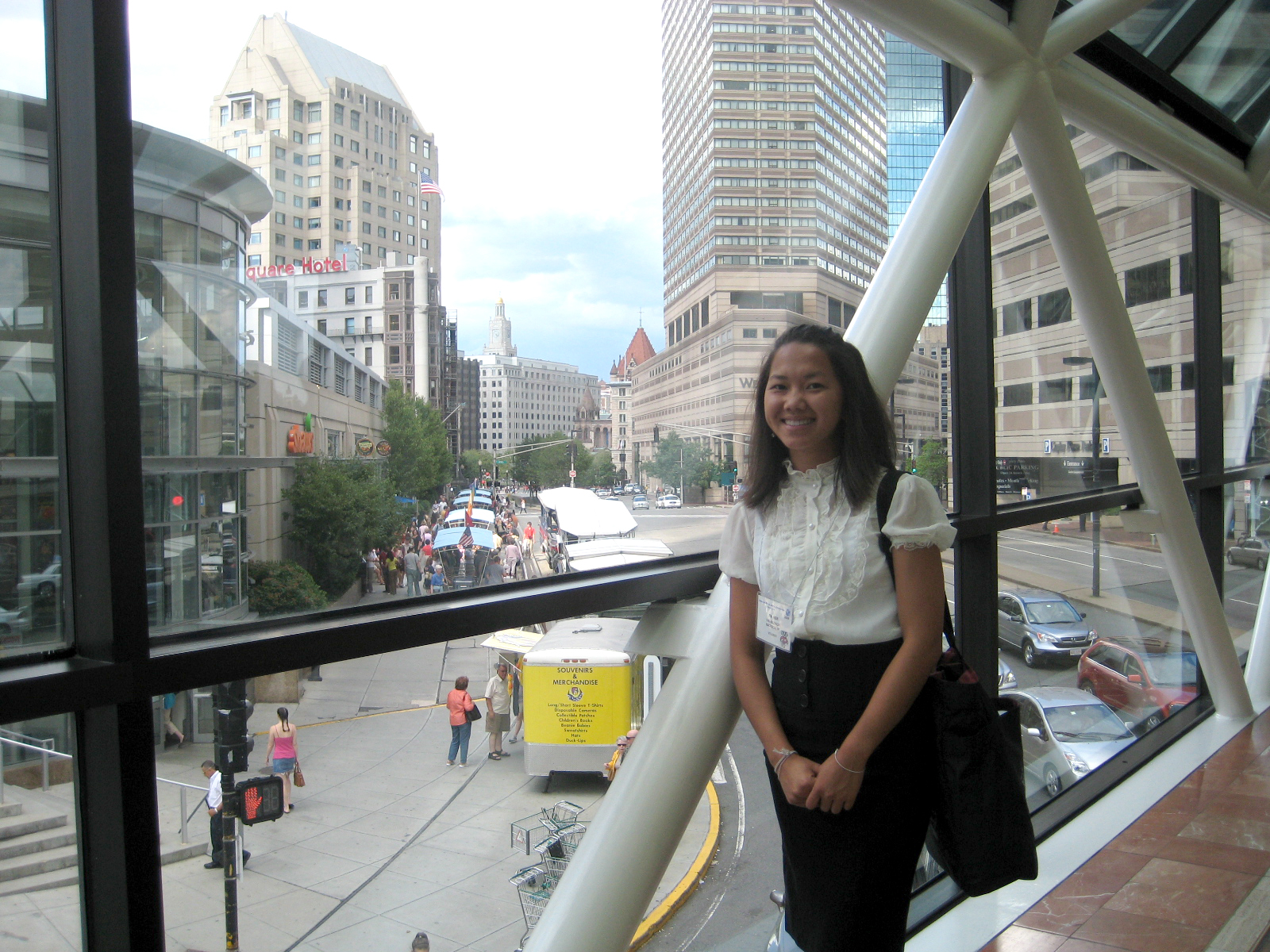Pa Her smiling while standing on a skybridge in Boston. The large windows of the skybridge reveal several pedestrians, food trucks, traffic, and large buildings.